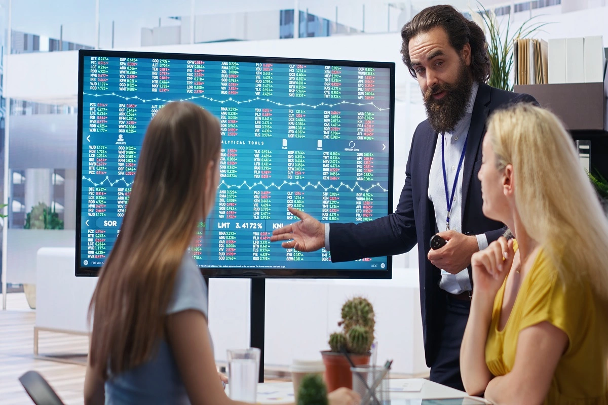 Business presenter explaining stock market and financial data on a large screen to two clients in a modern office.